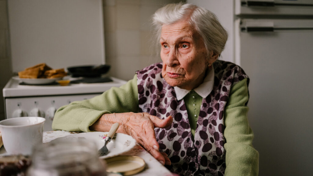older woman sitting at her kitchen table looking grumpy